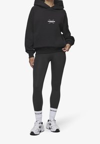 Sudadera negra con capucha, bolsillo frontal y logo blanco, combinada con leggings negros y zapatillas deportivas blancas con detalles en negro.
