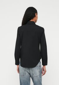 Melnas ar garām piedurknēm krekls ar apkakli, apaļām apakšmalām un pogām uz aprocēm, izgatavots no gludas auduma. Valdīts ar gaiši mazgātiem džinsa džinsiem.