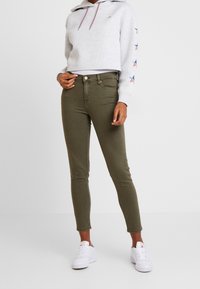 Persona che indossa un hoodie cropped grigio chiaro con disegni a stelle sulle maniche, pantaloni skinny verde oliva e sneaker bianche, in piedi contro uno sfondo semplice.