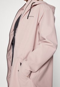 Veste imperméable rose clair avec capuche, fermeture éclair à l'avant, poignets élastiques et poches latérales. Dotée d'un logo sur la partie supérieure gauche.