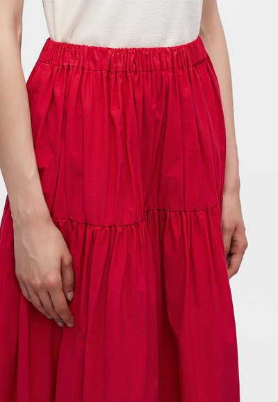 Femme portant une jupe rouge vif froncée avec une taille élastique et un haut blanc, la main reposant doucement à son côté.