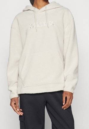 Person iført en cremefarvet, tekstureret fleece-hoodie med "SOMEWHERE" broderet på fronten og sorte bukser med sidelomme til bagage.