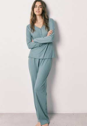 Ung kvinde stående barfodet iført lyseblå ribbet langærmet top og matchende løse bukser mod en ensfarvet hvid væg.