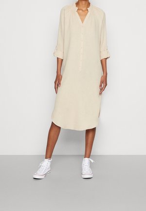 Kvinde iført løs, langærmet beige kjole med knapper og opsmøgede manchetter samt hvide høje sneakers, stående på en grå gulv.