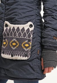 Vadderad marinblå jacka med en texturerad ficka i grått och vitt mönster med gula accenter, säkrad med svarta knappar.