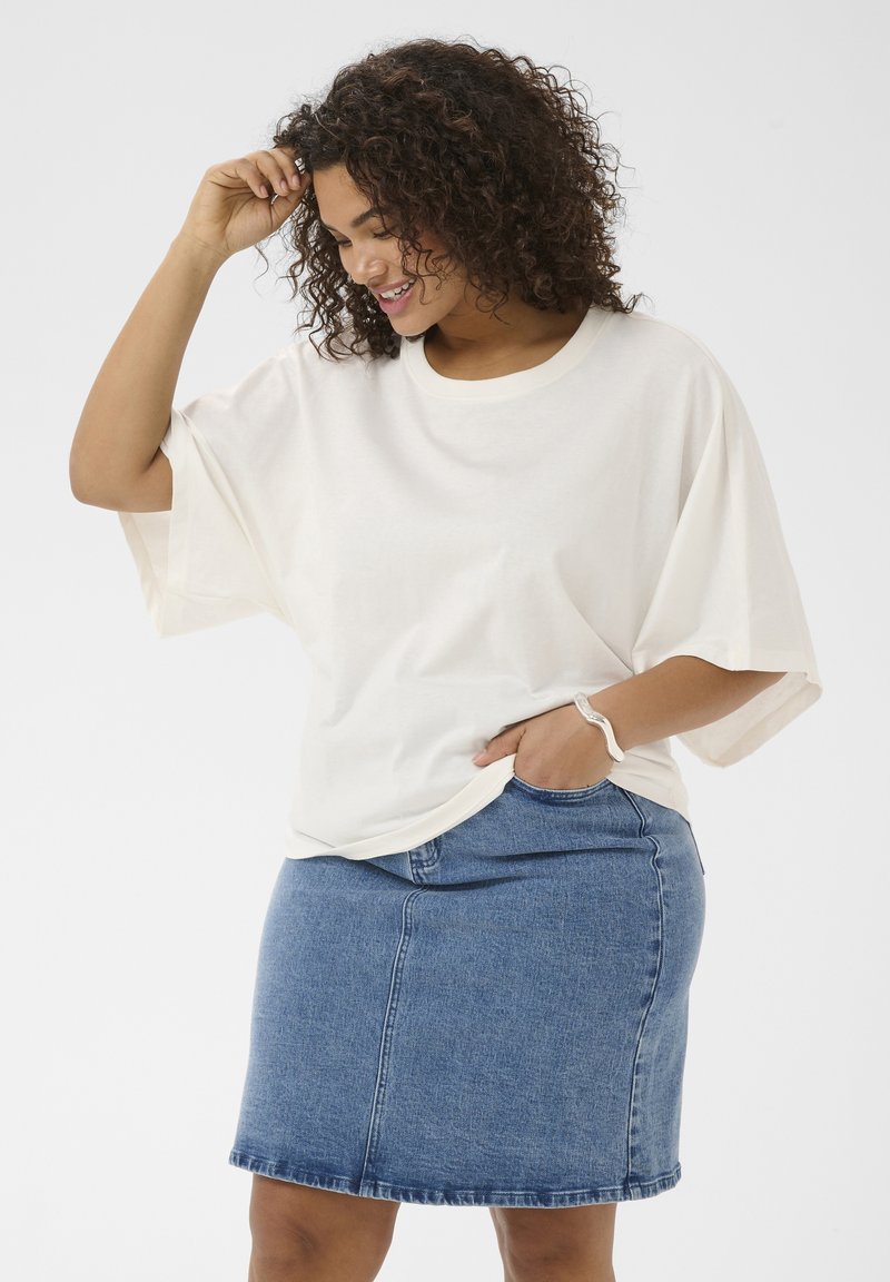 Femme aux cheveux bouclés portant un t-shirt blanc ample et une jupe en jean bleue, souriante et regardant vers le bas, main dans la poche de la jupe.