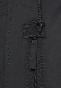 Tissu noir zippé, présentant un extérieur texturé avec une languette plate. La fermeture éclair est centrée et renforcée en haut.