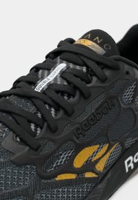 Sapatilha atlética preta com um cabedal de malha texturizada, sola de borracha, atacadores pretos grossos e detalhes amarelos com a marca "Reebok".