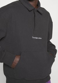 Black pullover sweatshirt with a polo collar, long sleeves, and front pocket. Features white "Calvin Klein Jeans" embroidery on the chest.