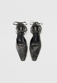 Chaussures à talons noires en cuir avec un bout pointu, un design entrecroisé et une finition texturée. Présentent un enroulement décoratif à la cheville avec des nœuds et des accents en perles.