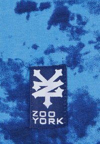 Blått stoff med et sprutmønster i mørkere nyanser. En rektangulær etikett med en hvit grafikk og teksten "ZOO YORK" er synlig.