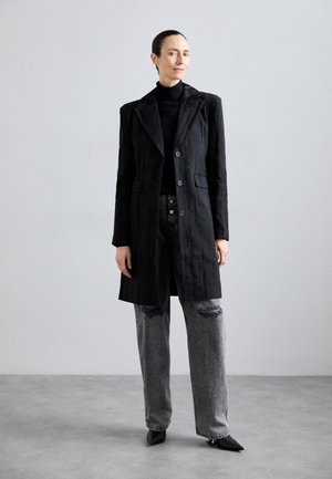 Schwarzer Texturmantel mit Reverskragen, Knopfverschluss und Fronttaschen, kombiniert mit abgewetzten grauen Jeans und spitzen schwarzen Schuhen.