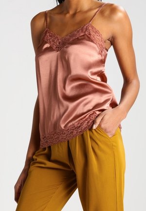 Kobieta w różowo-różowej satynowej bluzce na ramiączkach z koronkową lamówką i plisowanych spodniach w kolorze musztardowym, ręka w kieszeni na jednolitym tle.