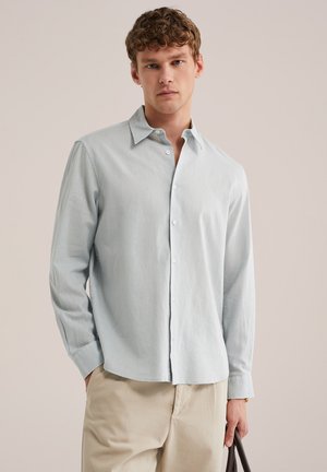 Jeune homme portant une chemise bleue claire à boutons et un pantalon beige, tenant une poignée de sac marron foncé sur un fond uni.