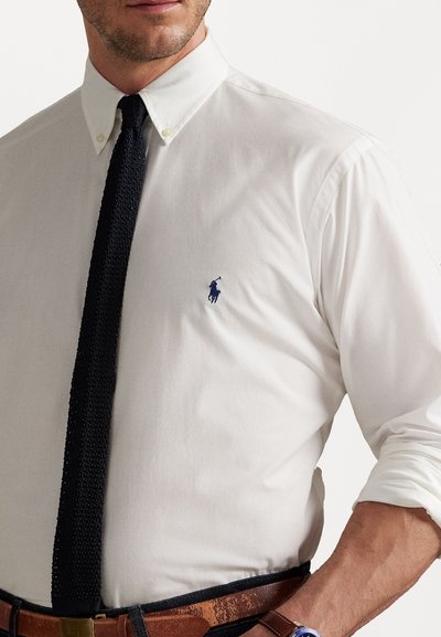Chemise blanche à boutons avec un logo bleu, cravate en tricot noire et ceinture marron. La chemise possède des manches retroussées et un design de col classique.