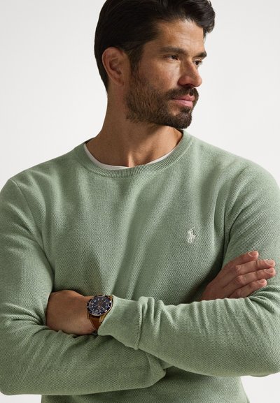 Knit sweater in light green, crew neck, textured fabric, featuring a small logo on the chest. Brown watch with a dark face on the wrist.