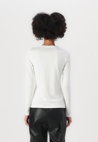 Camicia bianca a maniche lunghe con scollo rotondo, superficie liscia, design aderente, indossata con pantaloni neri a vita alta, vista da dietro.