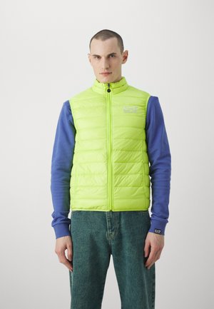 Zelena puffer brezrokavnik z zadrgo, ki ima šivano teksturo in detajl logotipa. Skombiniran z dolgimi rokavi modre majice in denimom.