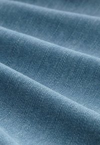Tela de mezclilla azul con una textura suave, plegada en suaves ondas que muestran sutiles variaciones en tonalidad y un patrón de tejido fino.