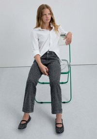 Camisa blanca de botones, jeans negros texturizados y bailarinas negras adornadas con tachuelas, sentada en una silla verde contra un fondo minimalista.