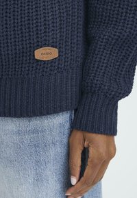 Marineblauwe trui met een textuur gebreid patroon, geribbelde manchetten en een tan leren label. Draagt over lichtdenim jeans, waarin handdetails zichtbaar zijn.
