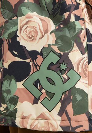 Grünes DC-Logo auf einem Hintergrund mit rosa und cremefarbenen Rosen sowie dunkelgrünen Blättern auf einem floral gemusterten Stoff.