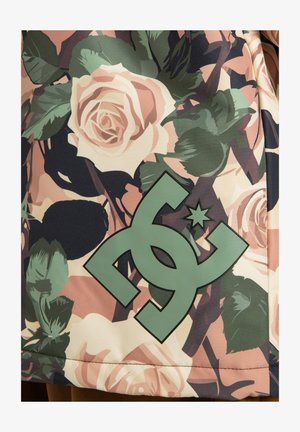 Grünes DC-Logo auf einem Hintergrund mit rosa und cremefarbenen Rosen sowie dunkelgrünen Blättern auf einem floral gemusterten Stoff.