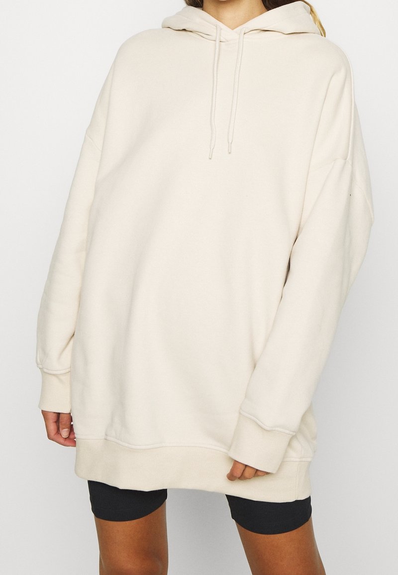 Sudadera oversized de color crema con capucha y cordón, puños acanalados y una textura suave y lisa. Combinada con pantalones cortos de ciclismo oscuros.