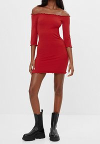 Červené šaty s odhalenými rameny, tříčtvrtečními rukávy, žebrovanou texturou a vroubkovanými okraji. Kombinované s černými robustními kotníkovými botami.