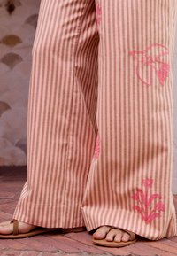 Pantalons rose à rayures évasées avec broderies florales roses. Le tissu semble léger, avec une texture lisse et une coupe décontractée. Sandales visibles.