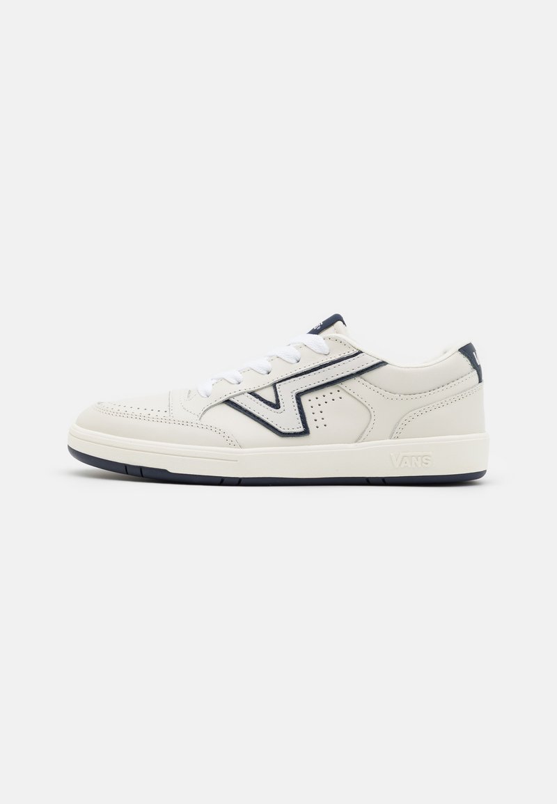 Baskets blanches basses avec une bande latérale en zigzag noire, des perforations, des lacets blancs et une semelle épaisse sur fond blanc uni.