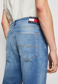 Jeans de mezclilla azul claro con corte recto, costuras naranjas, dos bolsillos traseros y una etiqueta de marca con colores rojo, blanco y marino.