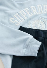 Ljusblå sweatshirt med vit broderad text och logotyp, i kombination med mörkblå joggers med resårmidja. Mjuk tygkvalitet och ribbade manschetter.