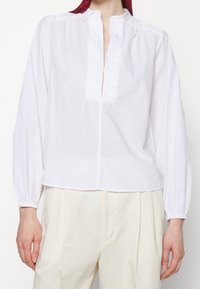 Valkoinen pitkähihaisen pusero, jossa on mandariinikaulus, ja siinä on poimutettua tekstiiliä edessä sekä joustavat hihansuut. Sileä, kevyt kangas.