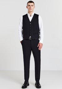 Schwarze maßgeschneiderte Weste mit fünf Knöpfen, getragen über einem weißen Hemd, kombiniert mit schwarzen Hosen und Schuhen. Strukturiertes Design mit Taschen.