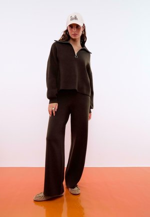 Maglione marrone con zip e pantaloni a gamba larga, realizzati in tessuto morbido. Cappello con la scritta "LA". Il modello indossa un anello di tendenza e scarpe marroni chiare.