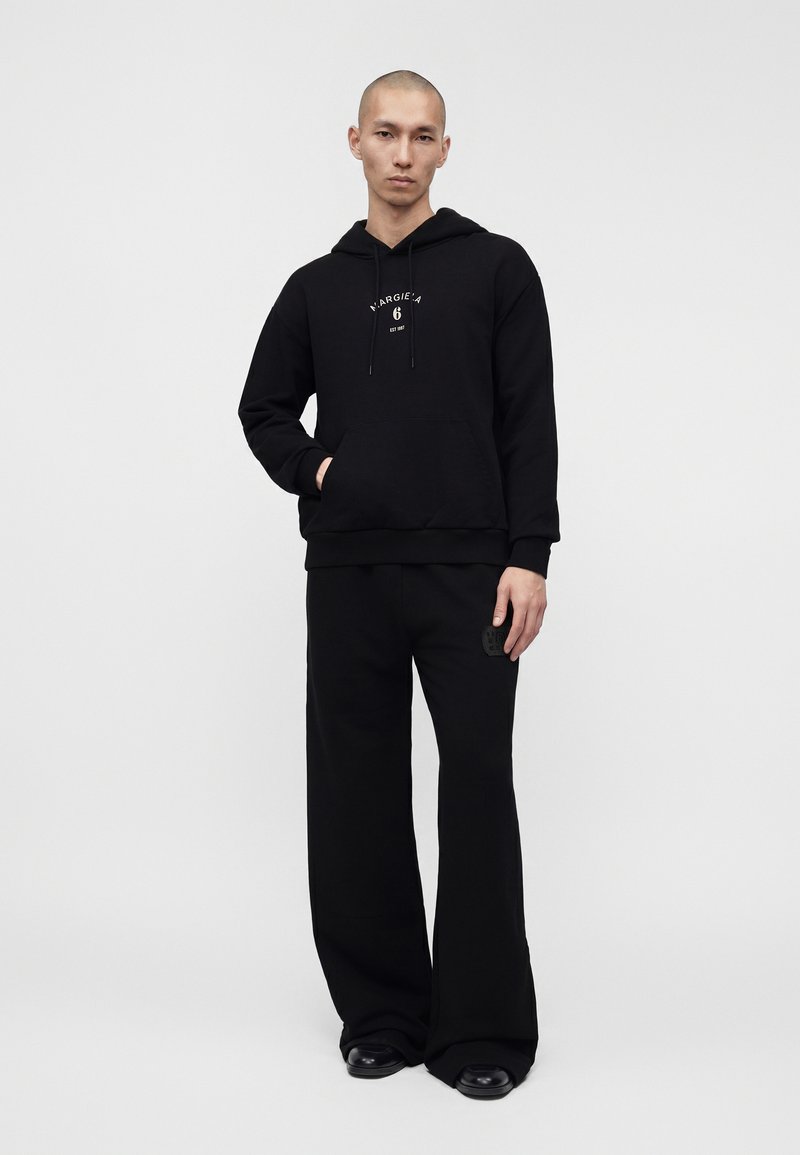Man staand, draagt een zwarte hoodie met voorzak en "MARGIELA 6" logo, wijde zwarte broek en zwarte schoenen tegen een effen achtergrond.
