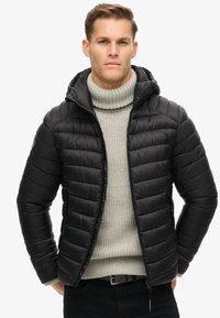 Chaqueta negra acolchada con capucha, cremallera frontal y bolsillos laterales; llevada sobre un suéter de cuello alto de canalé en gris claro. Textura suave, corte moderno.