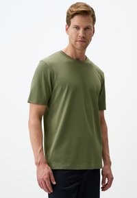 Oliwkowa bawełniana koszulka z krótkimi rękawami, okrągłym dekoltem i gładką fakturą. Zawiera subtelne detale szycia i luźny krój.