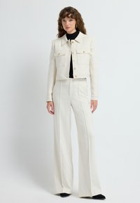 Afskåret cremefarvet blazer med guldknapper, parret med højtaljede, brede cremefarvede bukser. Sort turtleneck under. Glat tekstur.