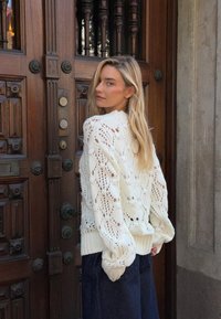 Valge lõngakootud pusa, millel on vabalt langev lõige, pitsmuster ja ribadest äärised, koos tumedate siniste laia säärega teksadega puust ukse taustal.