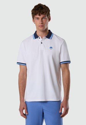 Uomo con polo bianca con colletto blu e righe sulle maniche, in piedi davanti a uno sfondo grigio chiaro uniforme.
