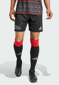 Pantalones cortos de fútbol negros y calcetines negros con puños rojos. La camiseta presenta un patrón geométrico y un logo. El calzado es negro y texturizado.
