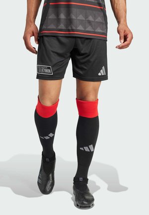 Sorte fodboldshorts og sorte sokker med røde manchetter. Trøjen har et geometrisk mønster og et logo. Fodtøjet er sort og tekstureret.