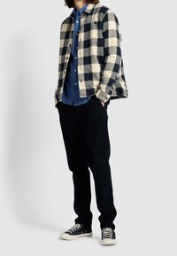 Camicia a scacchi in crema e blu marina; camicia blu sotto. Pantaloni scuri e sneaker nere completano l'outfit, caratterizzato da tessuti testurizzati.