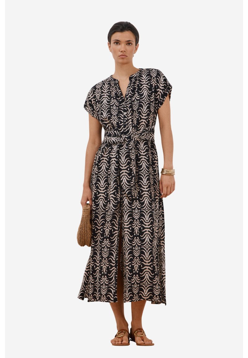 Maxikleid in Schwarz und Creme mit Wellenmuster, kurzen Ärmeln und tailliertem Schnitt. Kombiniert mit Sandalen und einer geflochtenen Handtasche.