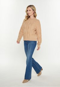 Beżowy sweter dzianinowy z teksturowanymi wzorami w romby i akcentami w postaci pomponów, zestawiony z dzwonowymi niebieskimi dżinsami oraz jasnobrązowymi szpilkami z ostro zakończonym noskiem.