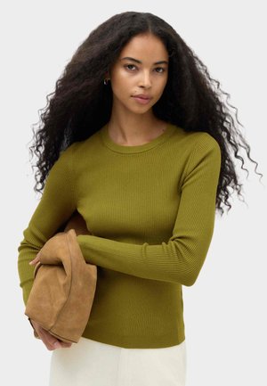 Vrouw met lang krullend haar, die een groene geribde trui draagt en een beige suède tas vasthoudt tegen een effen lichte achtergrond.