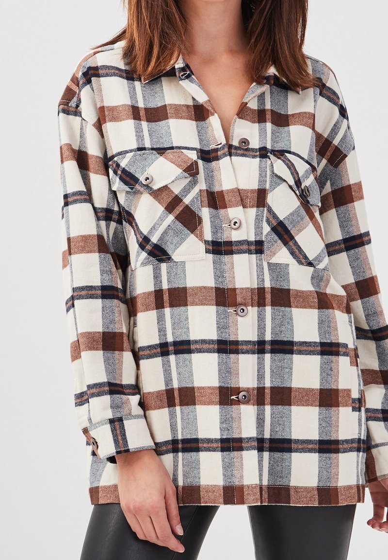Chemise en flanelle avec un motif écossais marron, beige et bleu. Elle a deux poches poitrine, des boutons sur le devant et des manches longues.