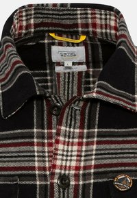 Camisa de franela a cuadros en rojo, negro y gris con un corte relajado, cuello con solapa, cierre de dos botones en la parte delantera y un parche circular con logo cerca del dobladillo.